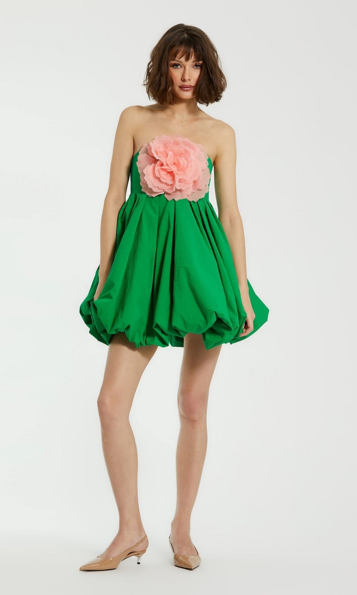 Wearing the Mac Duggal 20931 Short Bubble Party Dress in green with a large pink flower on the bodice and beige pointed-toe flats, a person stands against a plain white background.